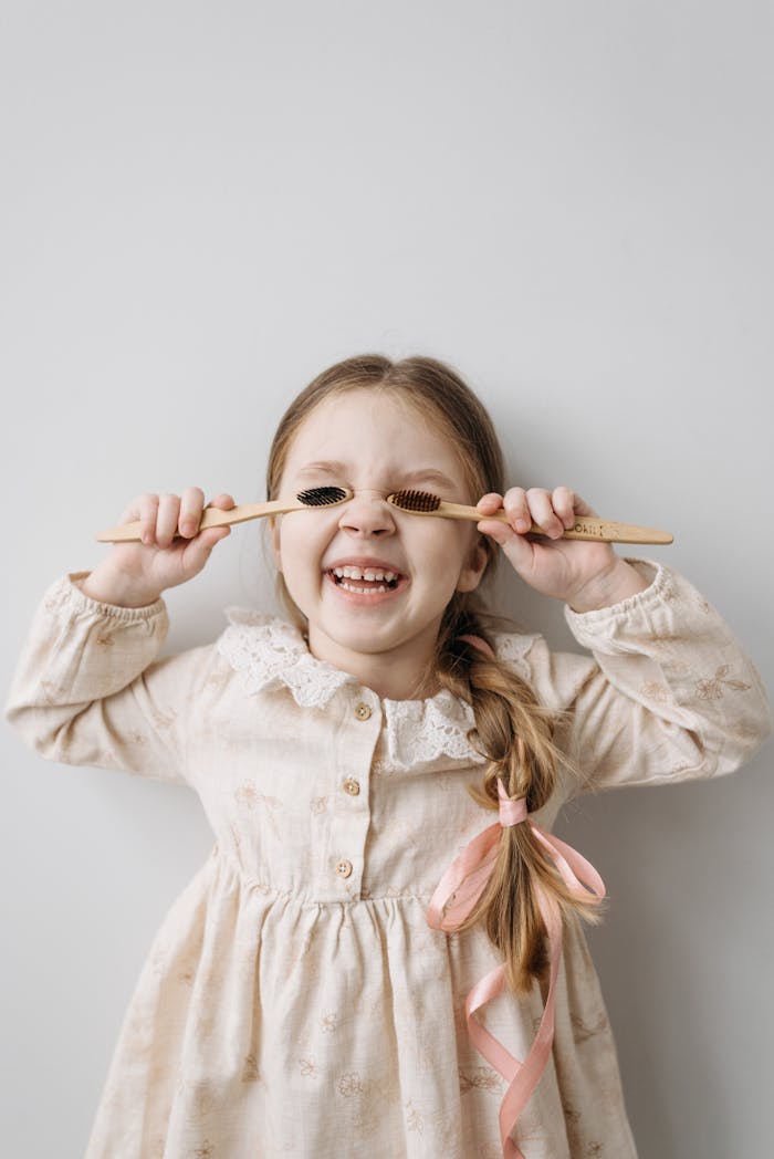 A happy child with braided hair playfully holding bamboo toothbrushes in front of her eyes.